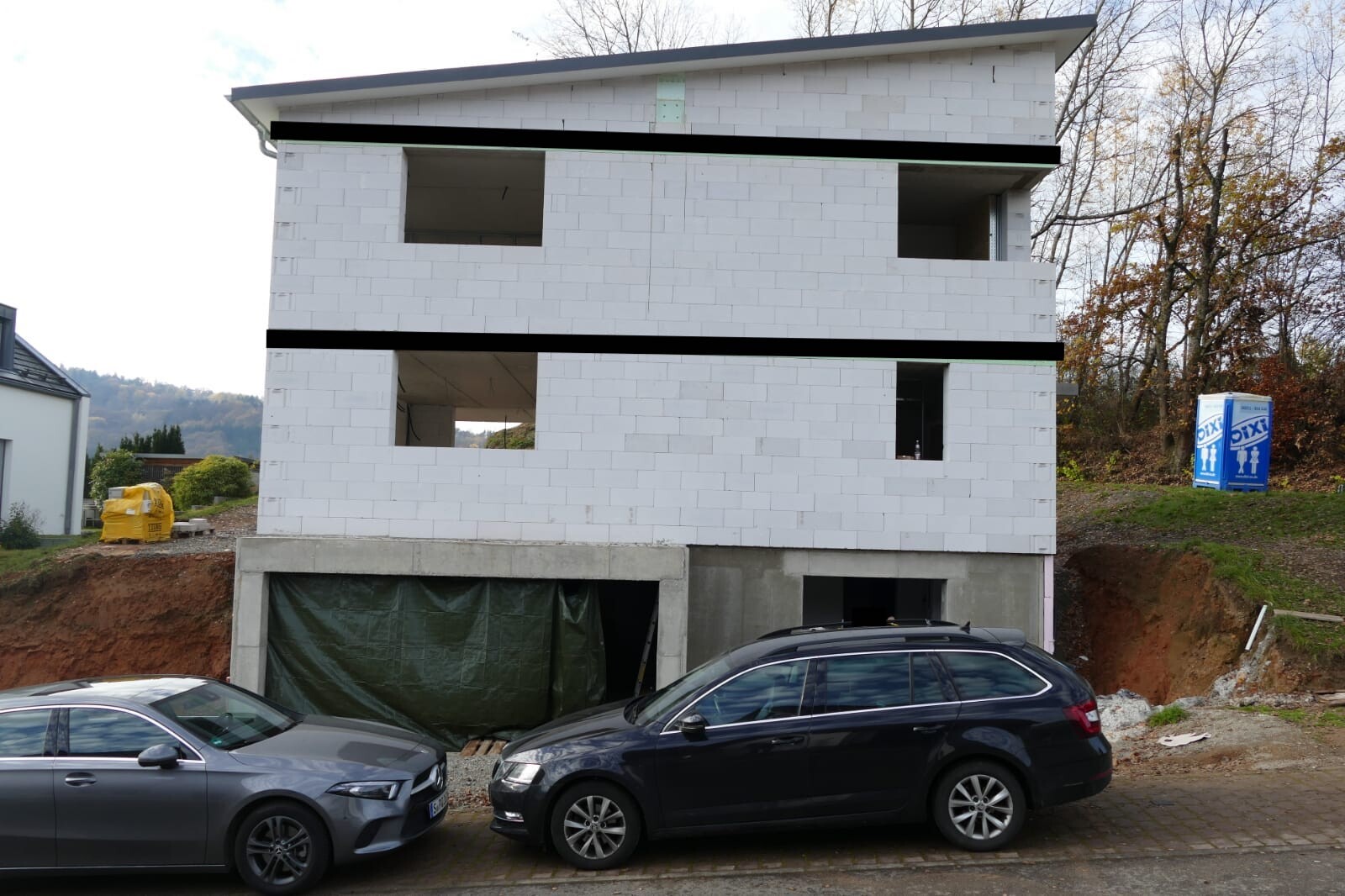 Dreistöckiger Neubau aus weißen Blöcken mit offenen Fenstern; vorn zwei Autos, Baucontainer und Toilette.