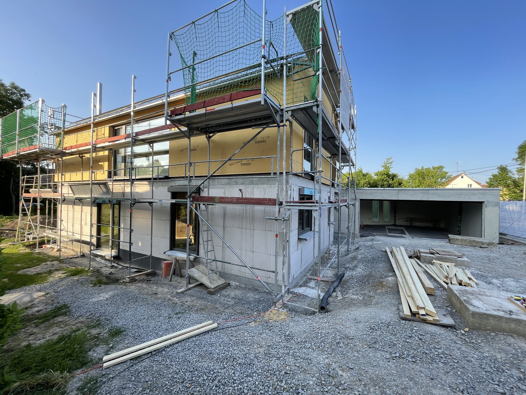 Zweistöckiges Haus im Rohbau mit Gerüst, grünem Sicherheitsnetz und Bauholz neben der Garage.