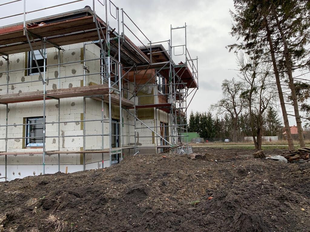 Baustelle am Rohbau eines Hauses mit Gerüst, Erde und umliegenden Bäumen.
