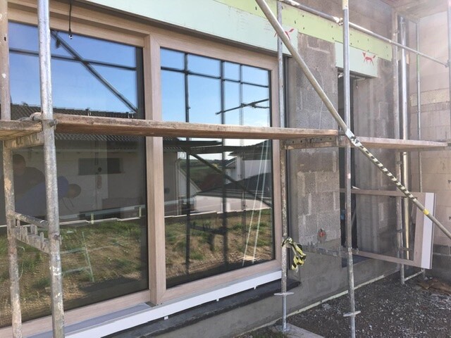 Großes Fenster mit Holzrahmen an einer Betonfassade, Baugerüst davor.