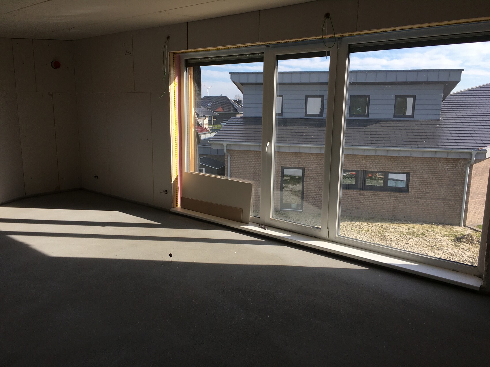 Unfertiger Innenraum mit großen Fenstern, Kabeln und Baustaub; Blick auf Nachbarhaus.