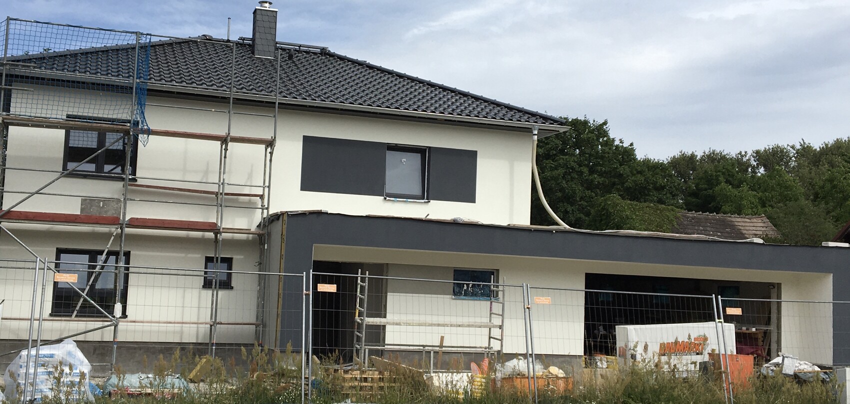 Moderne Hausbaustelle mit Gerüst, Bauzäunen und Baumaterialien vor weißer Fassade