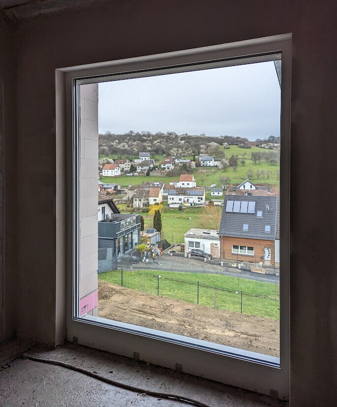 Blick aus unfertigem Innenraum durch ein großes Fenster auf Vorort mit Häusern, Feldern und Hügeln.