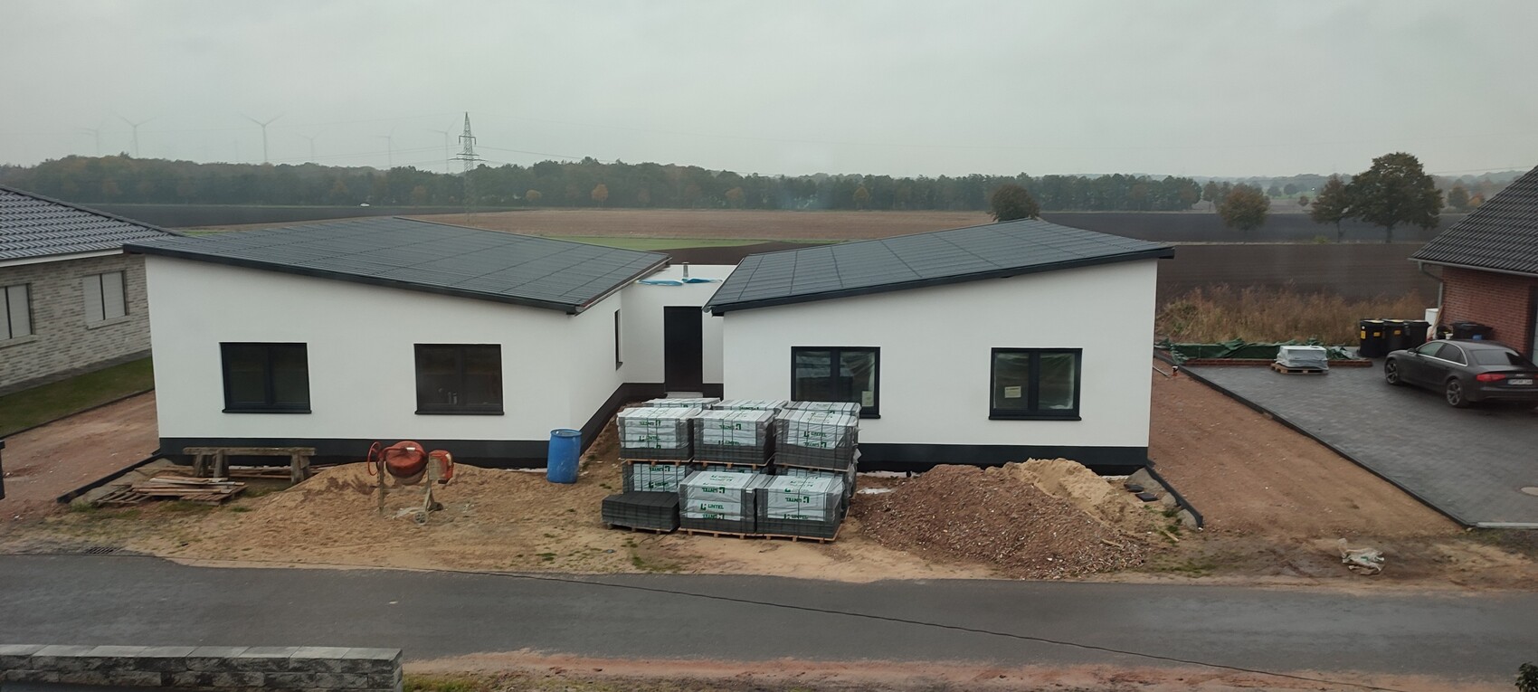 Neubau-Baustelle: zwei weiße Häuser, Paletten mit Material, Sandhaufen, Pkw, Felder mit Windrädern.