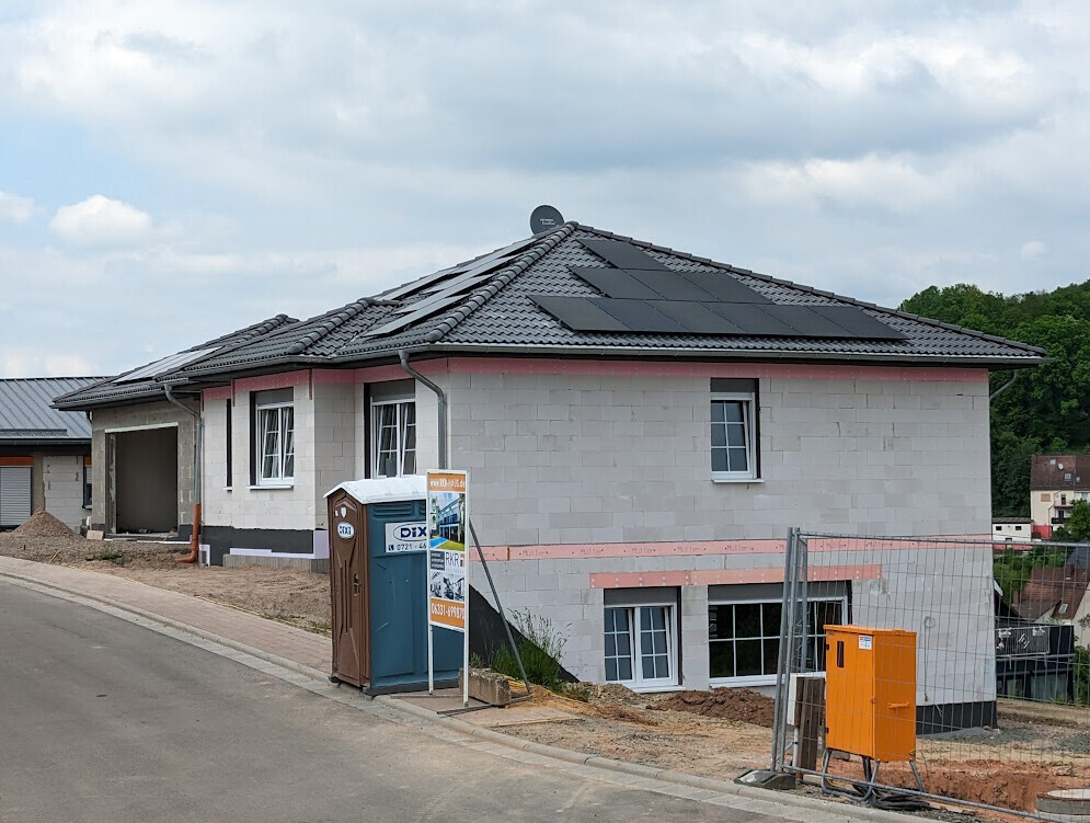 Neubauhaus mit Solarpanels, Bauzaun, Baucontainer und Toilettenhäuschen am Straßenrand