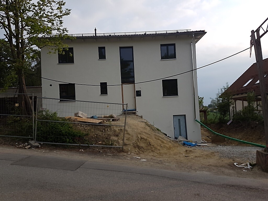 Mehrgeschossiges Haus im Rohbau auf Hang, Bauzäune, Baumaterialien vor Straße.