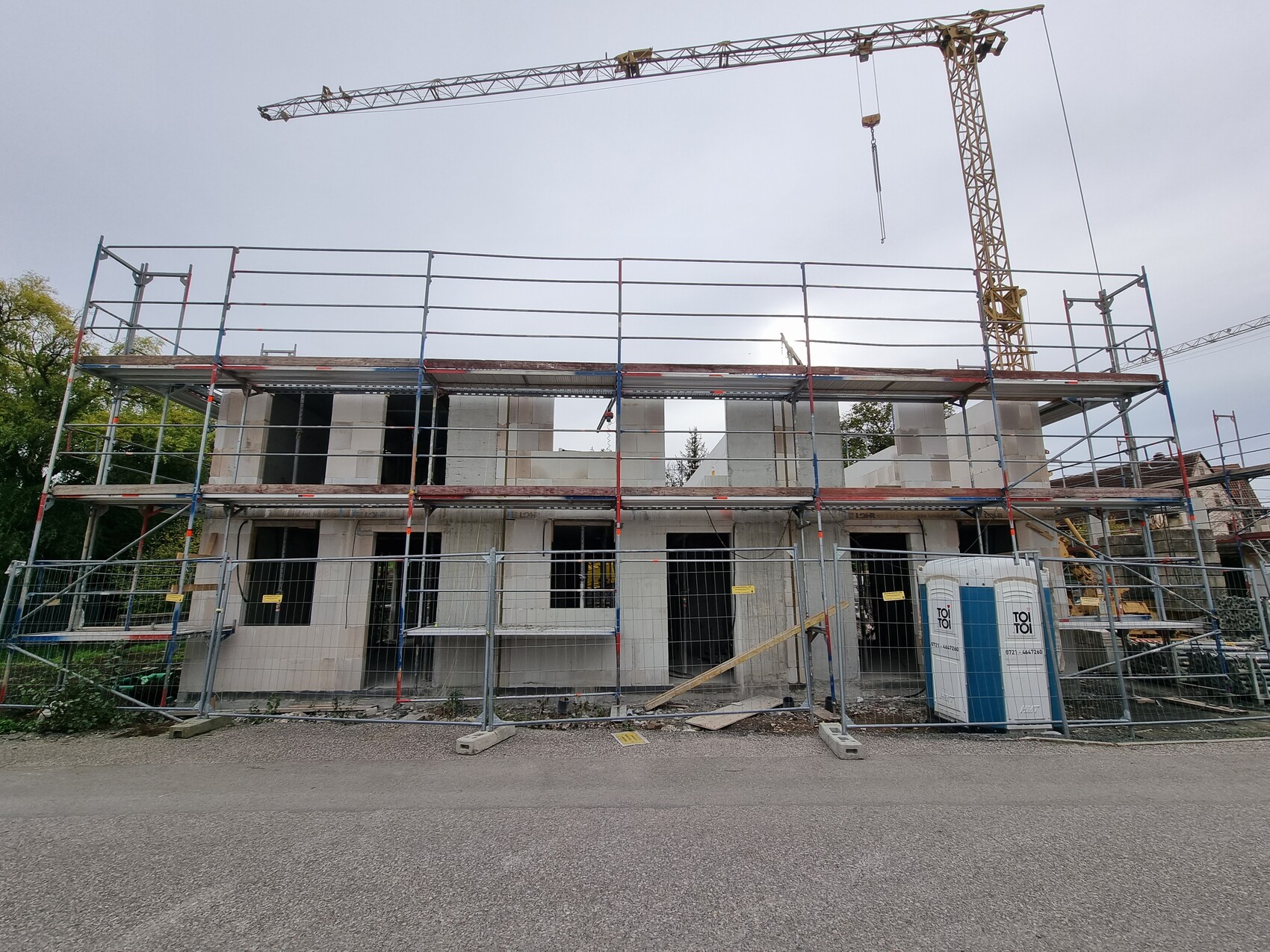 Baustelle mit Gerüst um zweistöckigen Rohbau, Kran im Hintergrund, Zaun und Toilettencontainer.