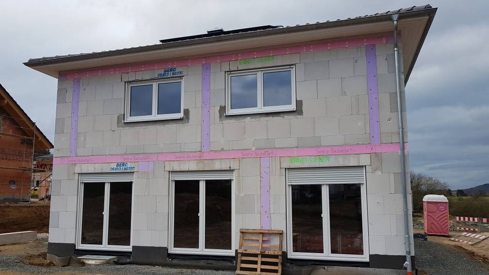 Zweistöckiges Rohbauhaus mit Fenstern, rosa Klebeband-Streifen und Baugeräten.