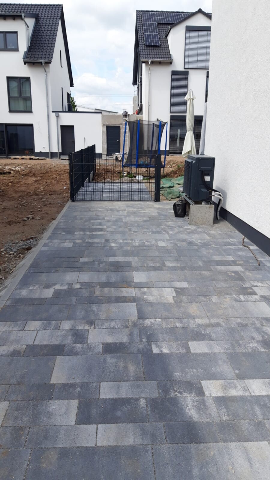 Gepflasterter Weg zum Hof mit Tor; Trampolin hinter Zaun, moderne Häuser mit Solarpanels.