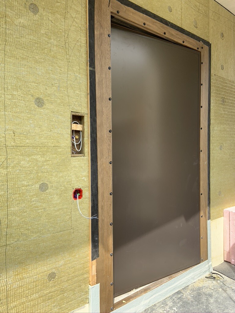 Große braune Metalltür in Holzrahmen vor gelb-grüner Dämmplatte; sichtbare Elektroleitungen in Wand.