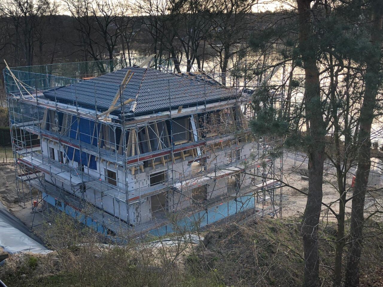 Mehrstöckiges Haus im Rohbau mit Gerüst ringsum, Dacharbeiten, Bauholz sichtbar, nahe Uferwald.