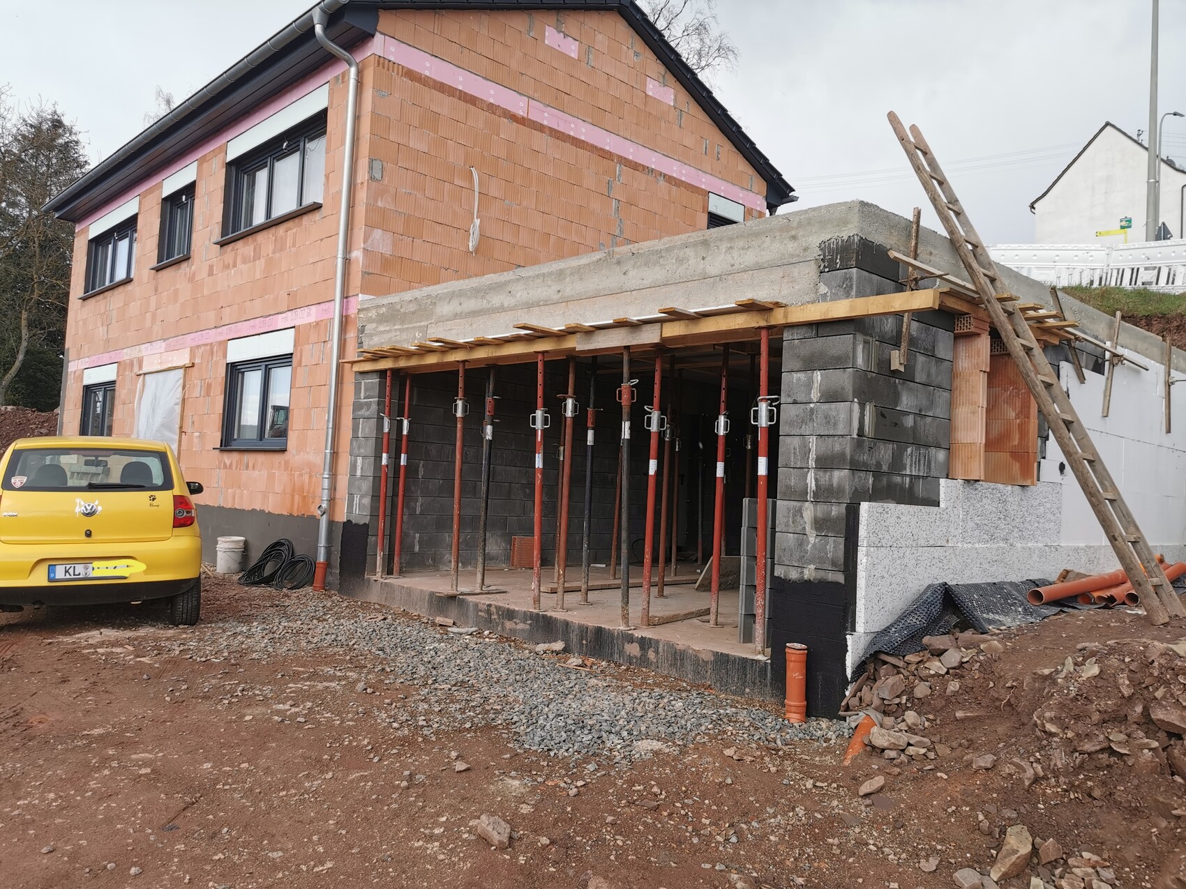 Unfertige Bauarbeiten an einem zweistöckigen Haus, Stützen, Holzbalken, Leiter; gelbes Auto davor.