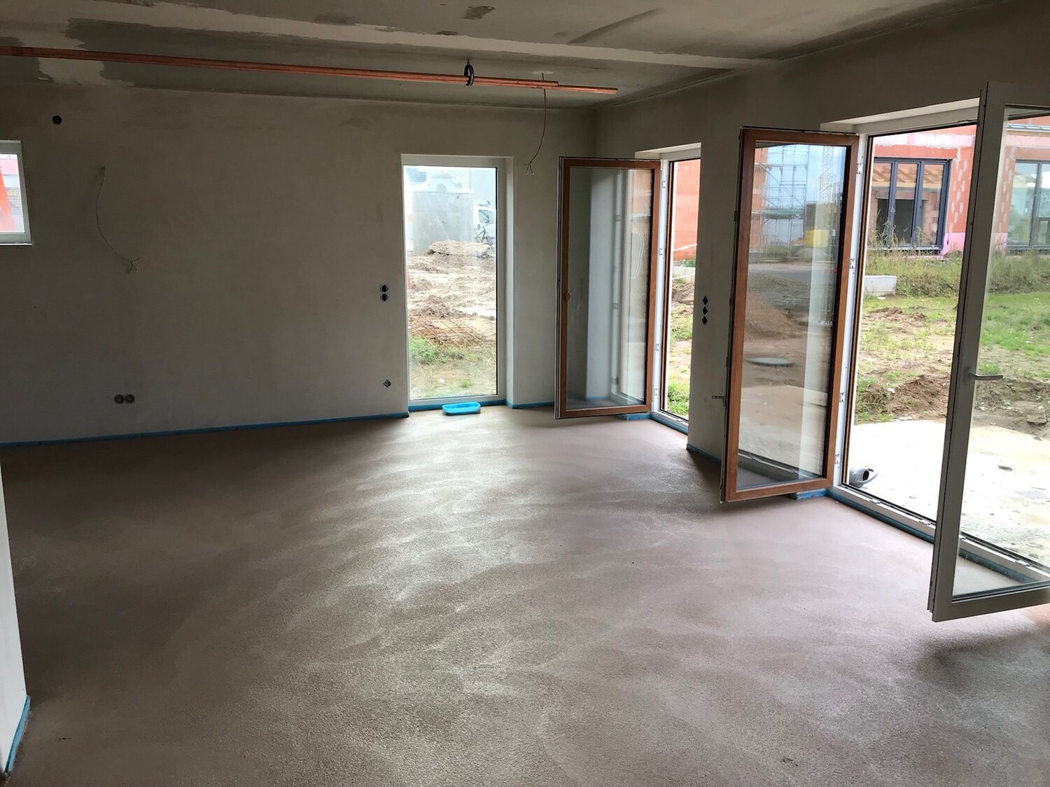 Unfertiger Innenraum mit großen Glas-Schiebetüren nach draußen; Baustelle und Erdarbeiten sichtbar.
