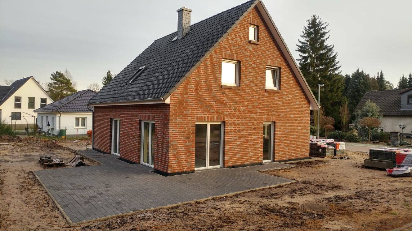 Rotes Backstein-Einfamilienhaus mit dunklem Ziegeldach und gepflasterter Terrasse; Baustellenszene.