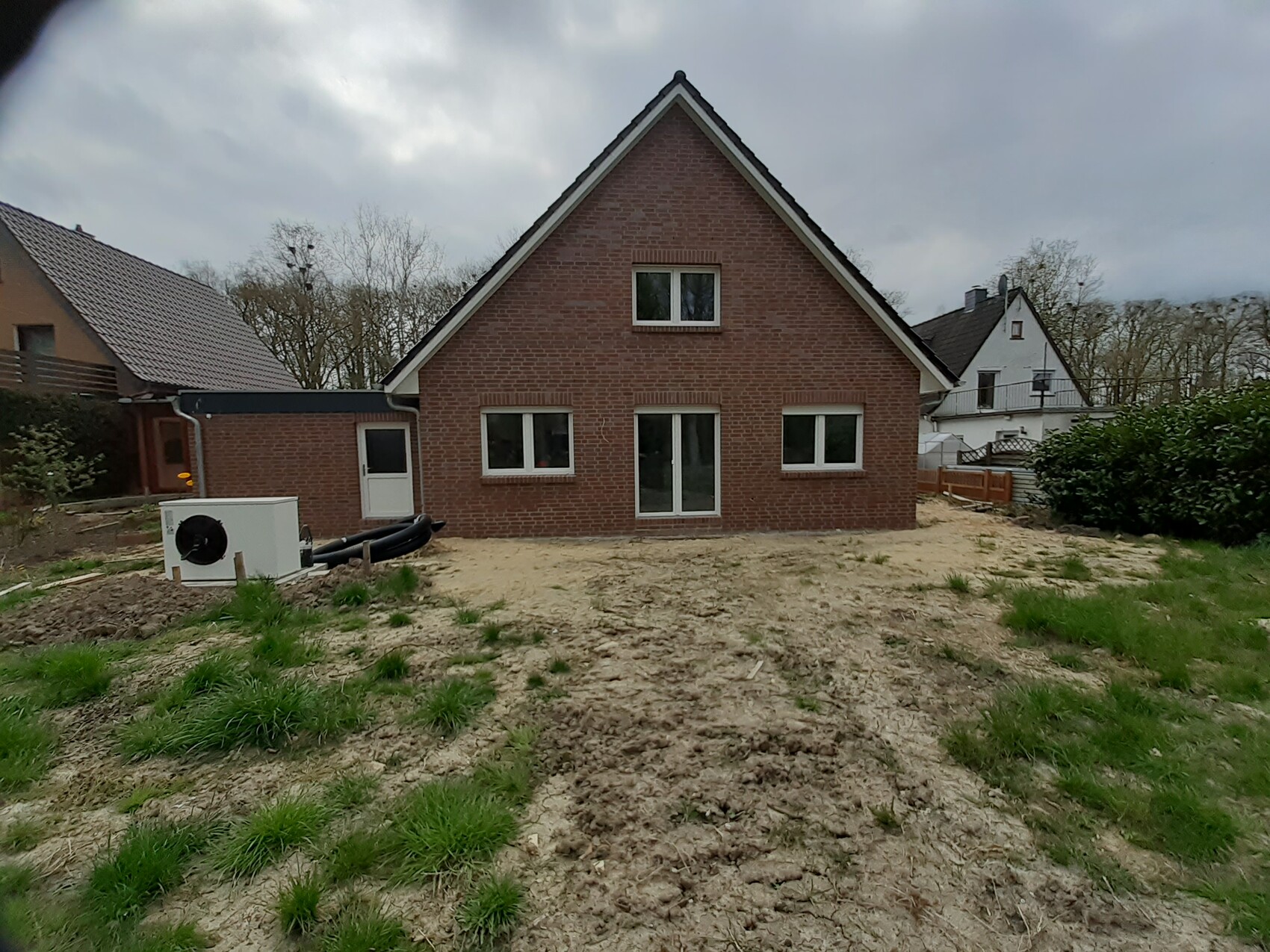 Neubau: rotes Backsteinhaus; unfertiger Garten mit weißer Außen-Wärmepumpen-Einheit und Rohren.