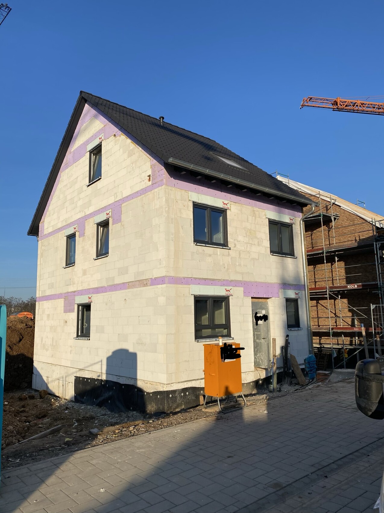 Einfamilienhaus im Rohbau mit hellen Steinblöcken, pinkfarbene Dämmung, Kran, Dach und Baugerüst.