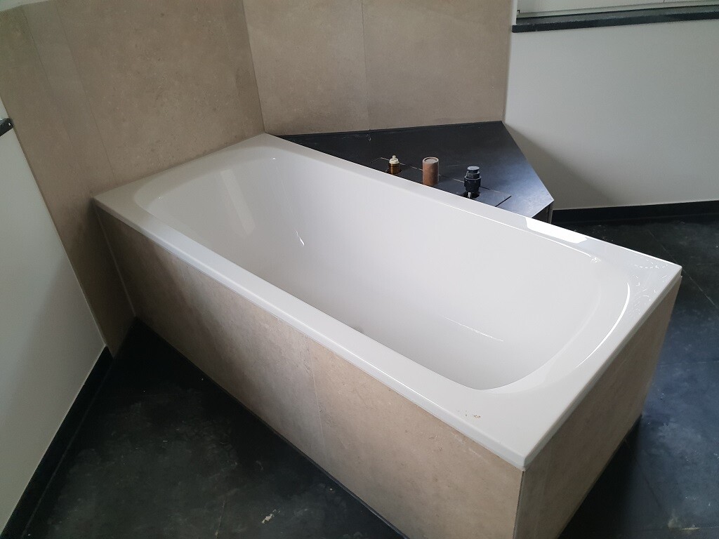 Weiße rechteckige Badewanne in beige gefliestem Bad, dunkle Arbeitsplatte mit Wasserleitungen.