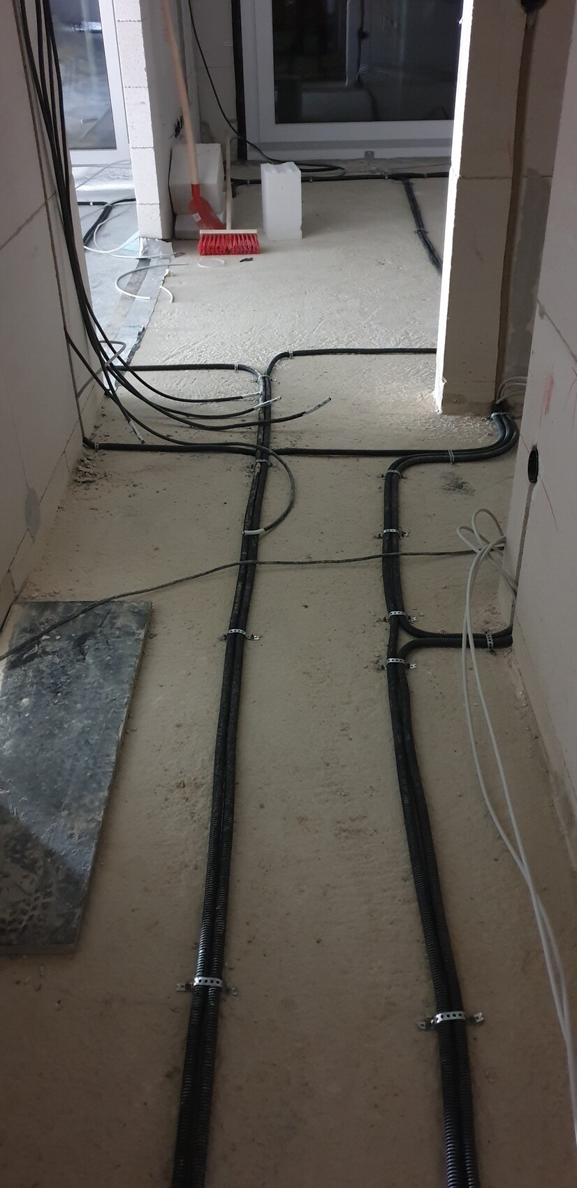 Baustellenflur mit schwarzen Kabelkanälen auf dem Boden, Putzreste und Reinigungswerkzeug.