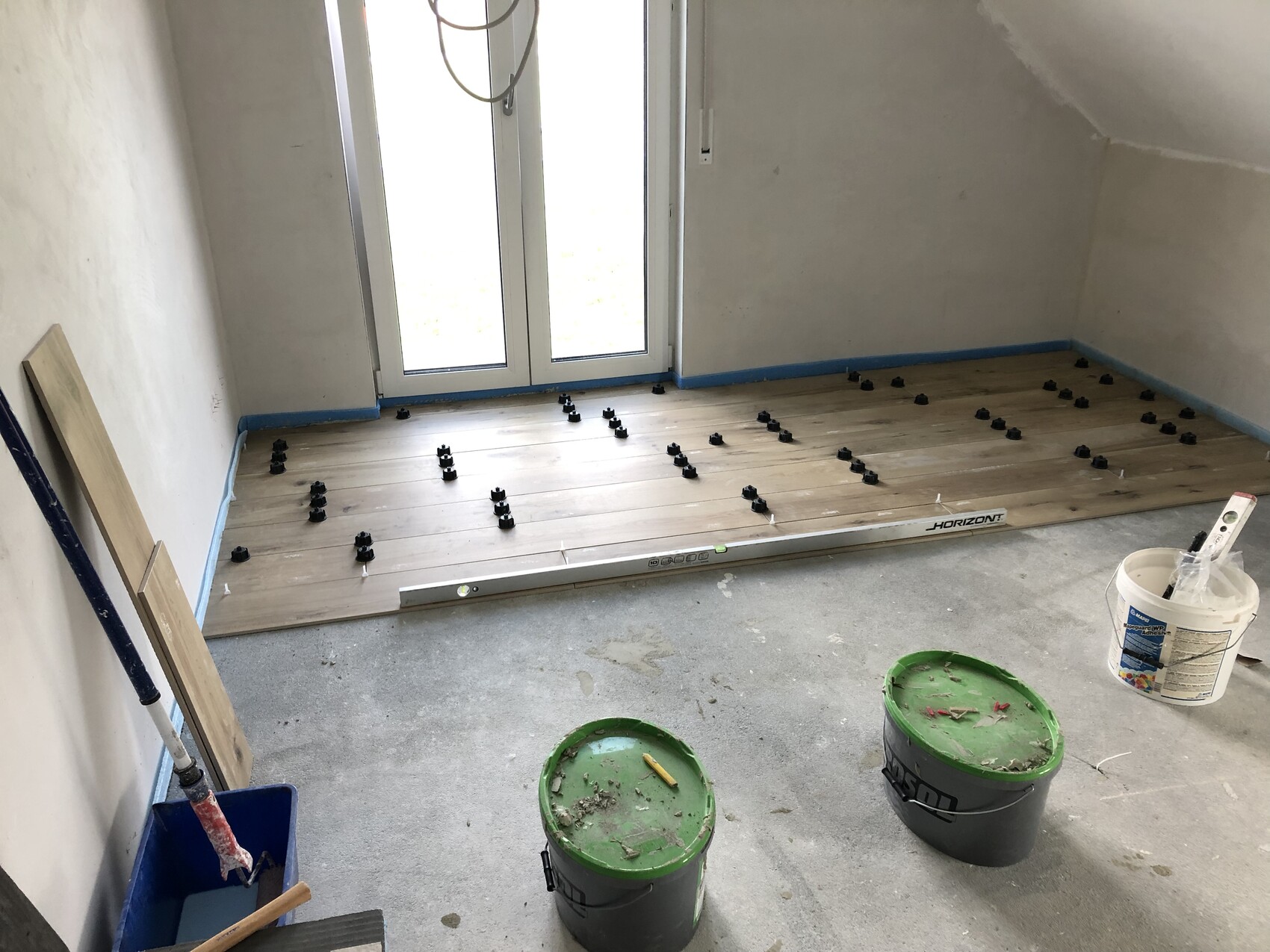 Raum mit Holzboden-Verlegung: Abstandhalter, Level, Eimer und Werkzeuge; Tür nach draußen.