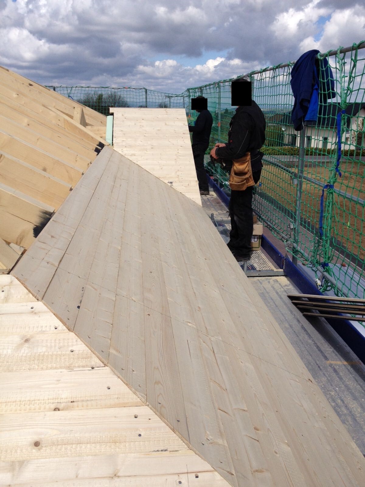 Zwei Bauarbeiter montieren Holz-Dachplatten auf einer Baustelle, grüne Schutznetze am Rand.