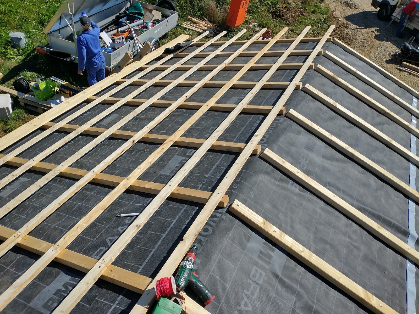Dachlatten liegen quer auf schwarzer Dachfolie; Bauarbeiter im Hintergrund.
