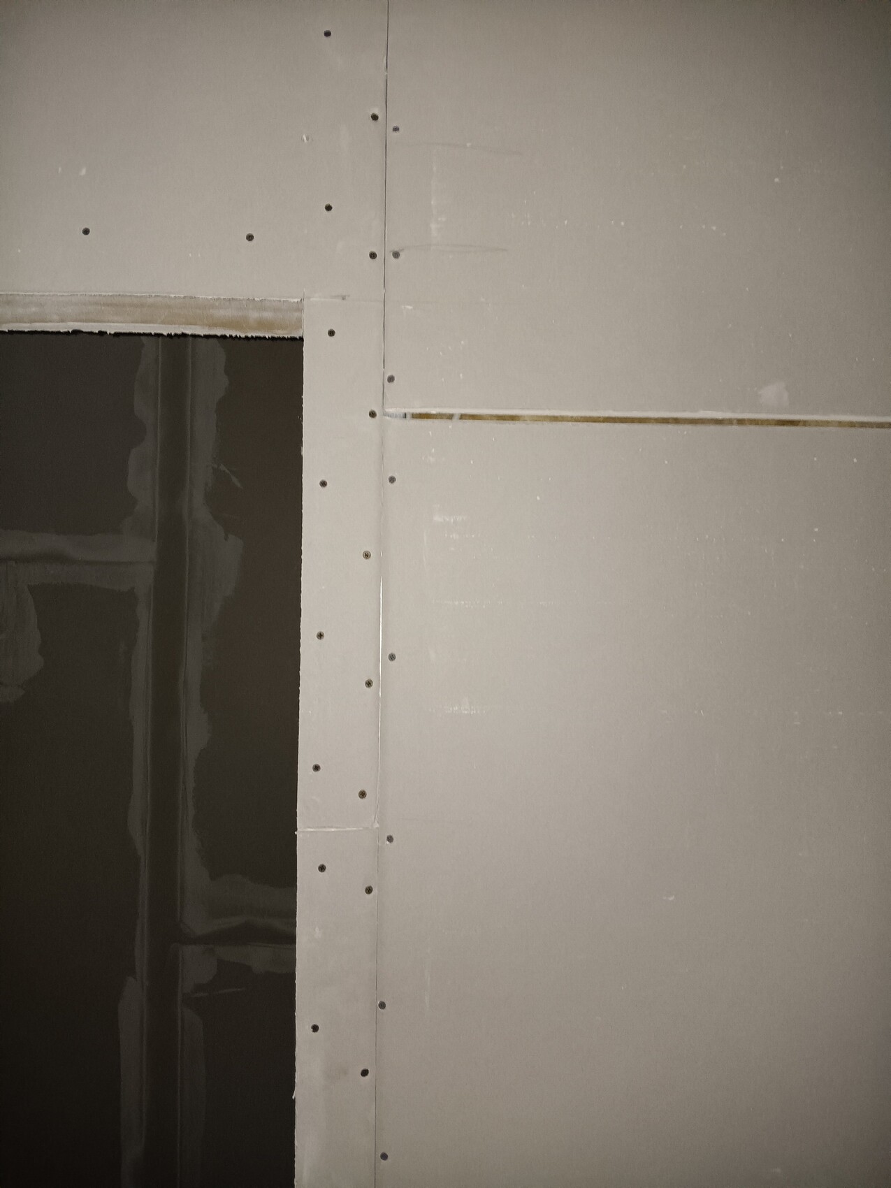 Eine Wand aus Gipskartonplatten mit Schrauben an den Fugen; offener dunkler Durchgang links.