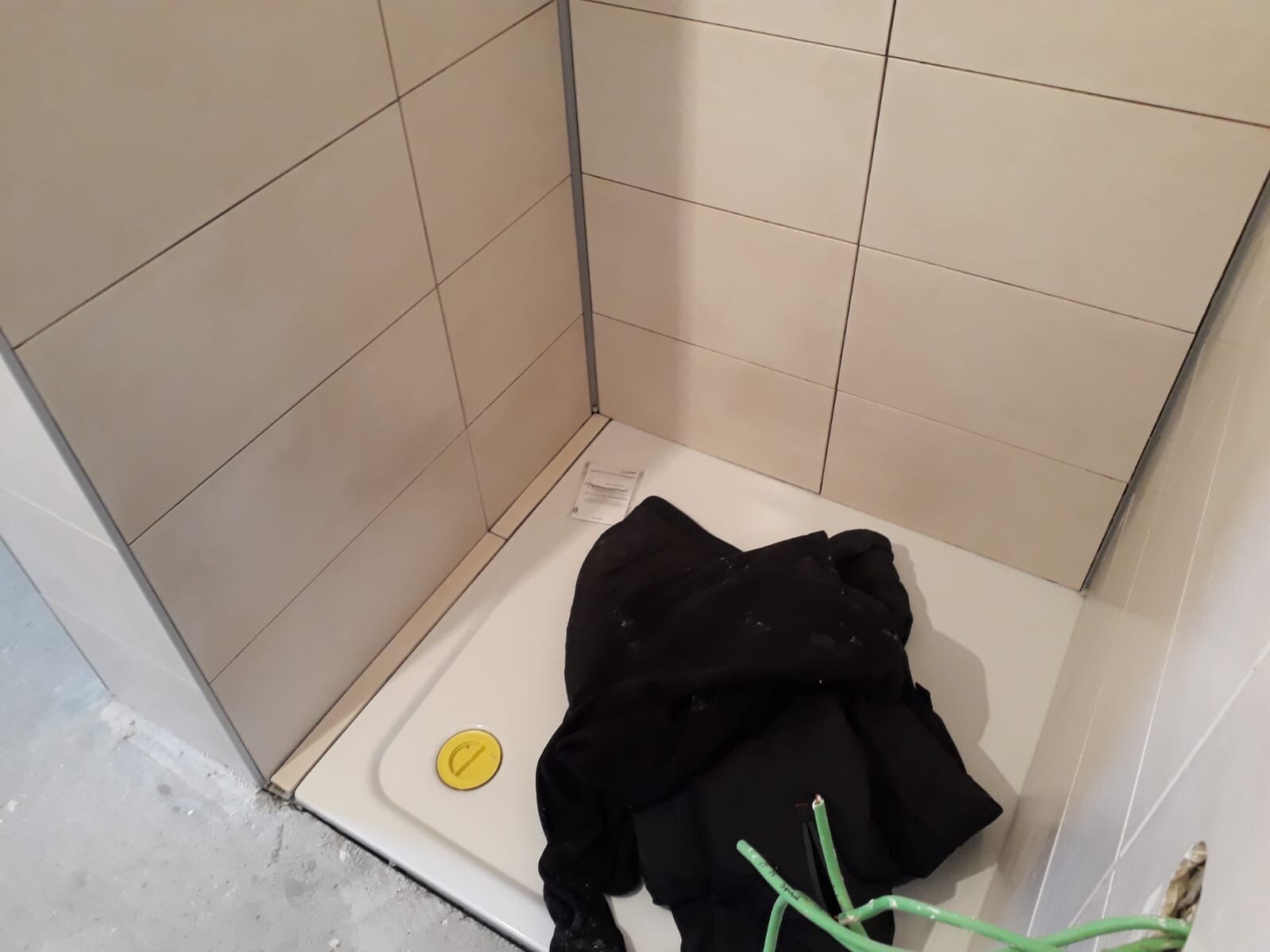 Dusche in Bau: beige Fliesen, weiße Duschwanne, gelber Abflussdeckel, schwarze Jacke.