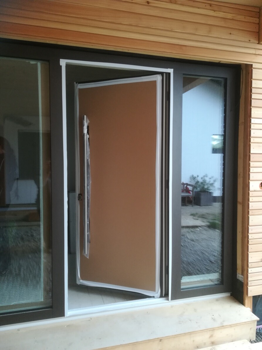 Braune Haustür hängt schräg im geöffneten Rahmen einer Holzfassade neben Glasfenstern.