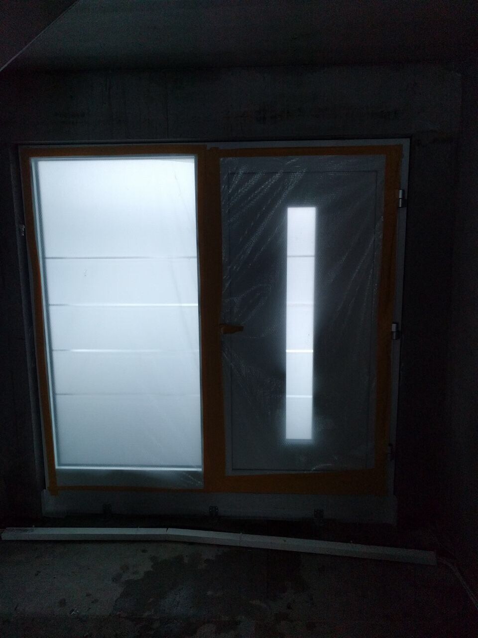 Neue Eingangstuer mit Holzrahmen und Glaseinsatz in dunklem Bauinneren, Schutzfolie sichtbar