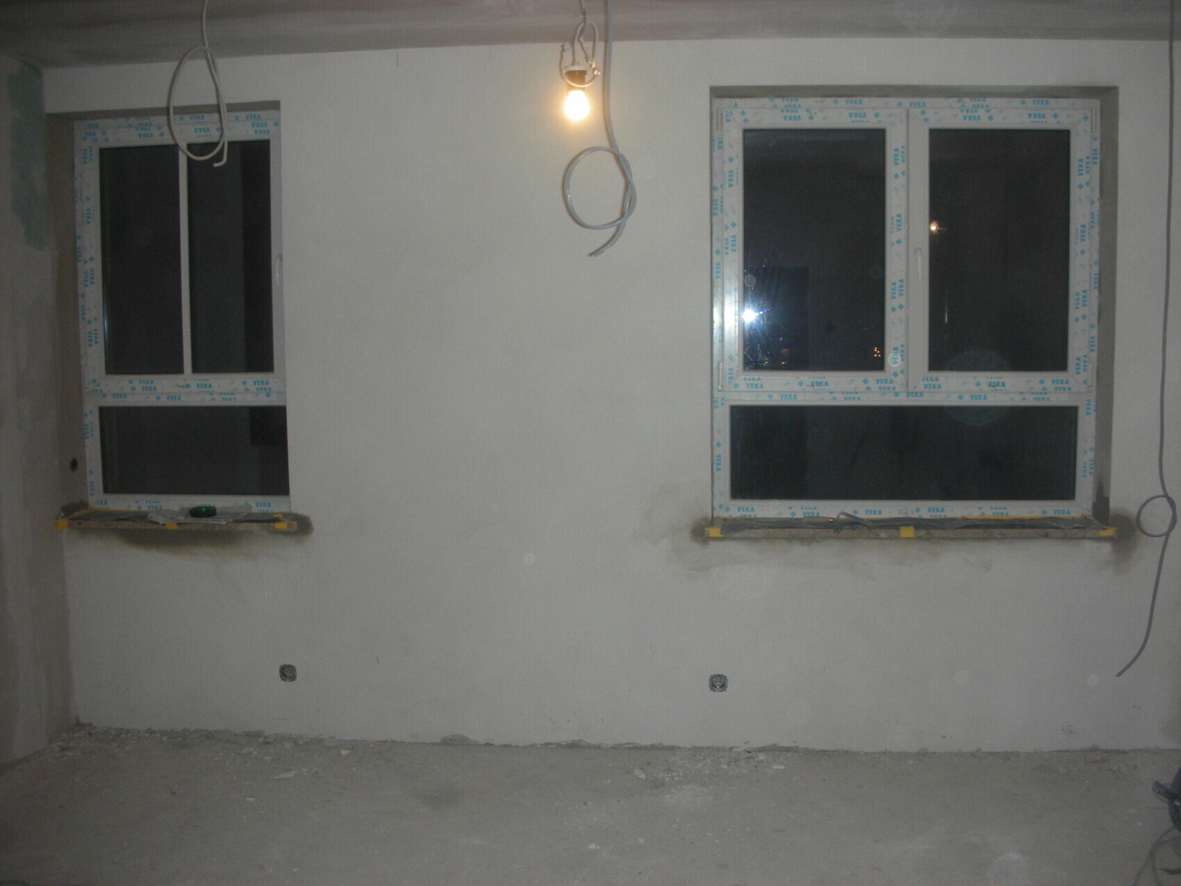 Baustelle: zwei Fenster, lose Kabel, hängende Glühbirne, staubiger Boden
