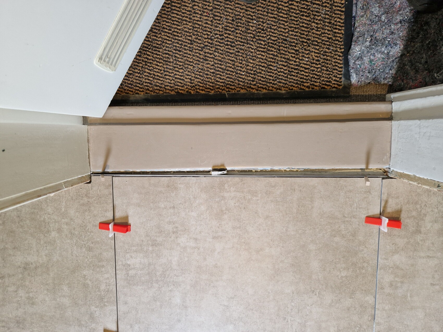Türschwelle zwischen beige Fliesen, rote Fliesenspacer an den Fugen, oben eine Teppichmatte.
