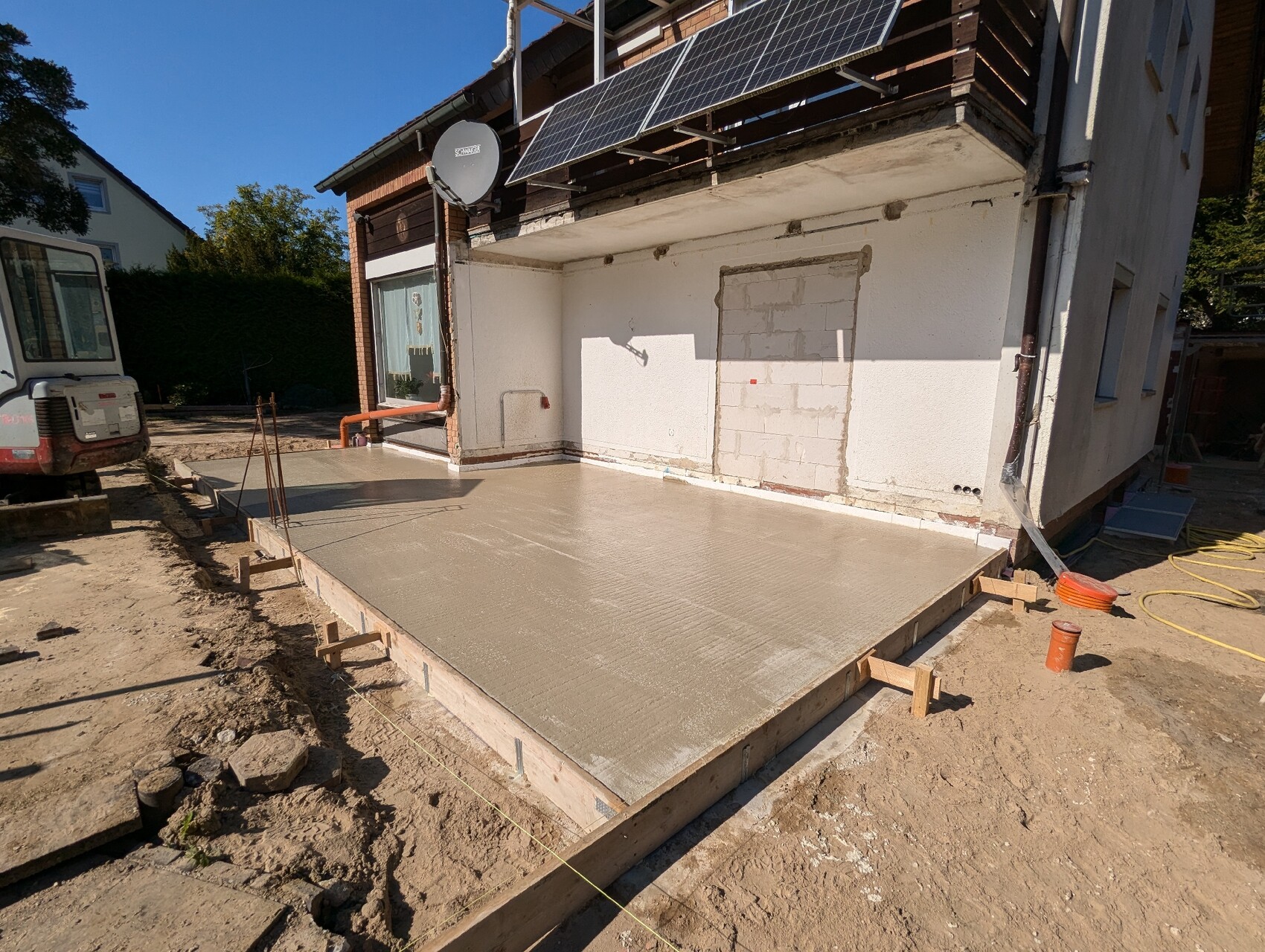 Frisch gegossene Betonplatte vor Wohnhaus; Baugeräte, Satellitenschüssel, Solaranlage auf dem Dach.