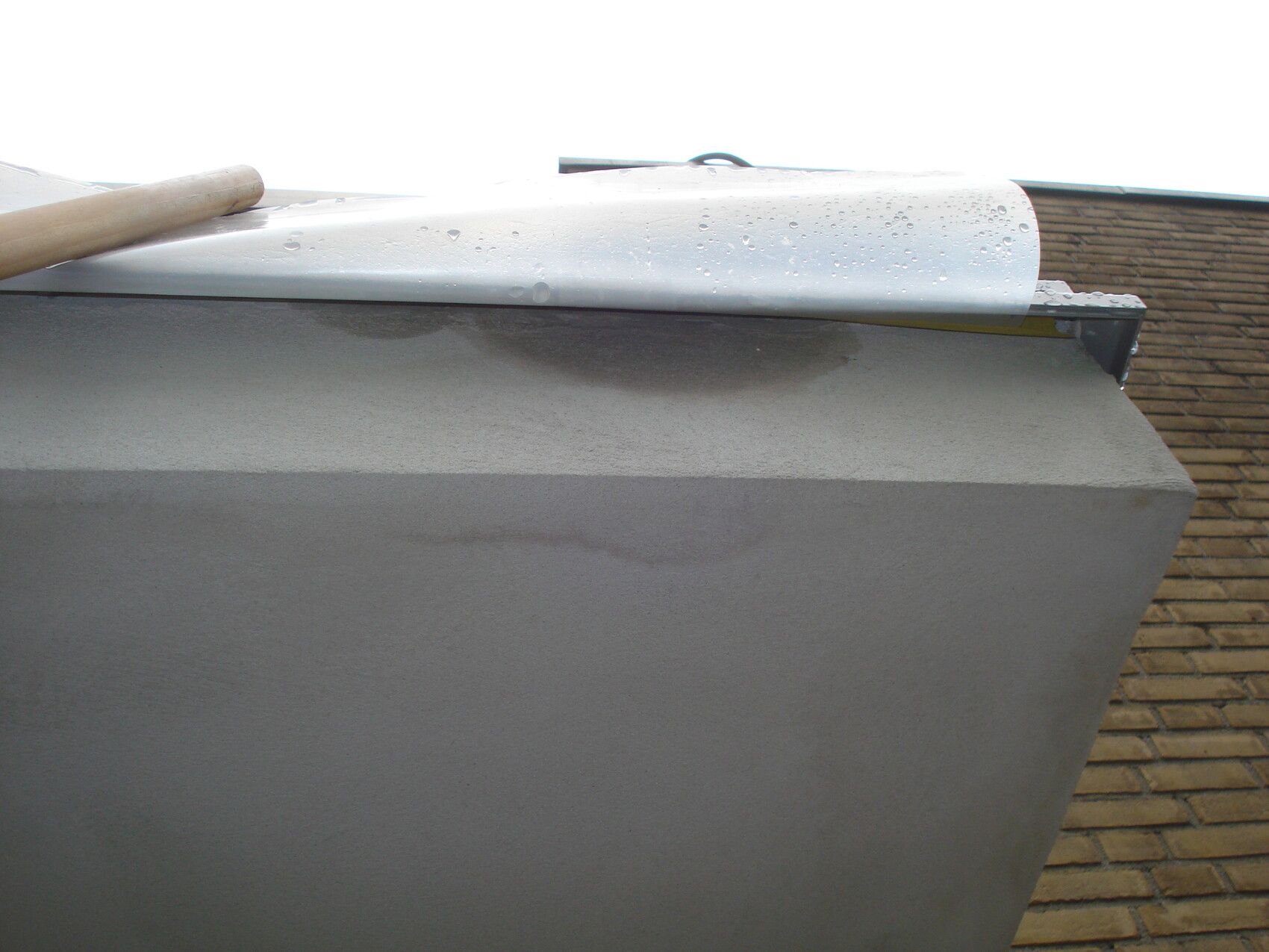 Nahaufnahme eines weißen Rollblechs über einer grauen Betonwand, Holzstab darauf; Regentropfen.