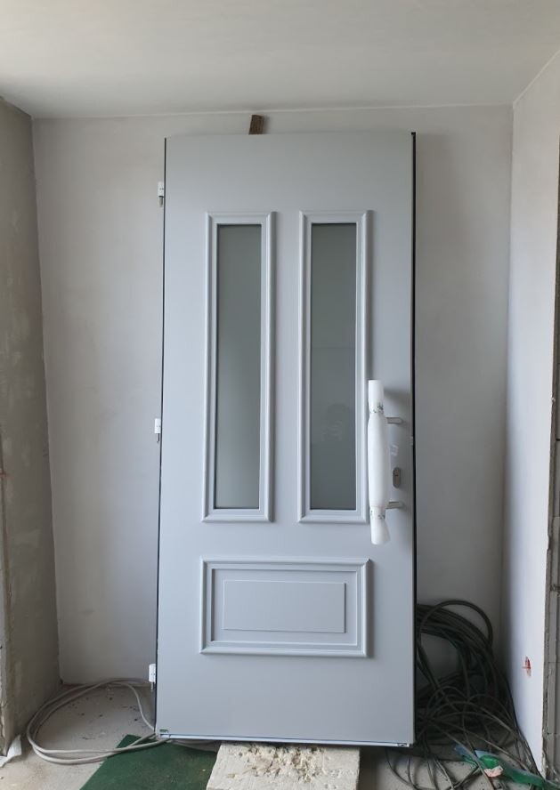 Weiße Haustür mit zwei schmalen Glasfenstern, Griff rechts, auf einer Baustelle.