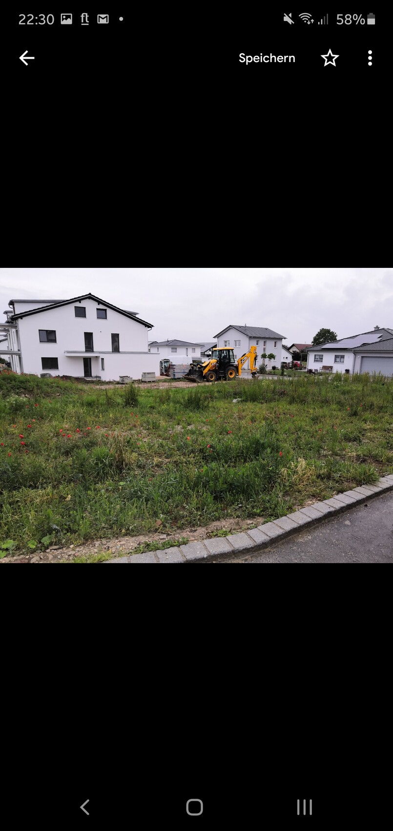 Gelber Bagger auf leerem Baugrund vor weißen Neubauhäusern in einer Wohnsiedlung; Gras im Vordergrund.