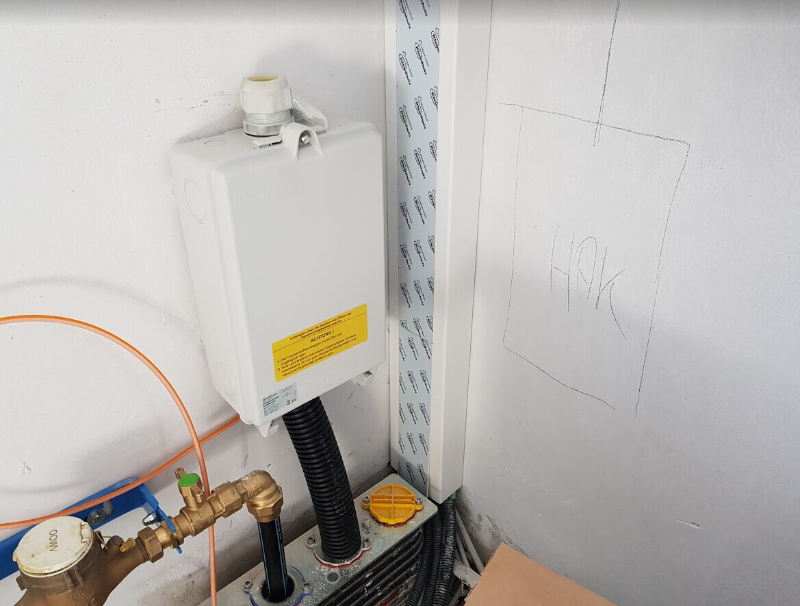 Weiße Wand-Steuerbox mit gelber Warnetikett, schwarzem Kabelschlauch und Kupferrohren.