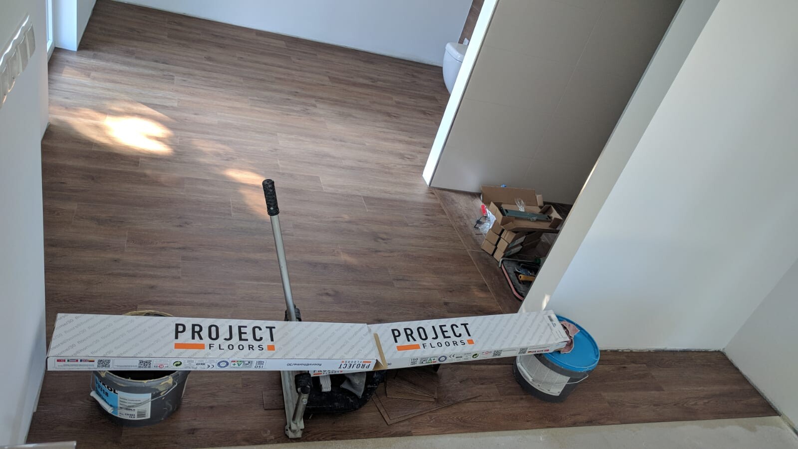 Renovierungsraum mit braunem Laminat, Verlegewerkzeug, Kartons und Eimer auf dem Boden.