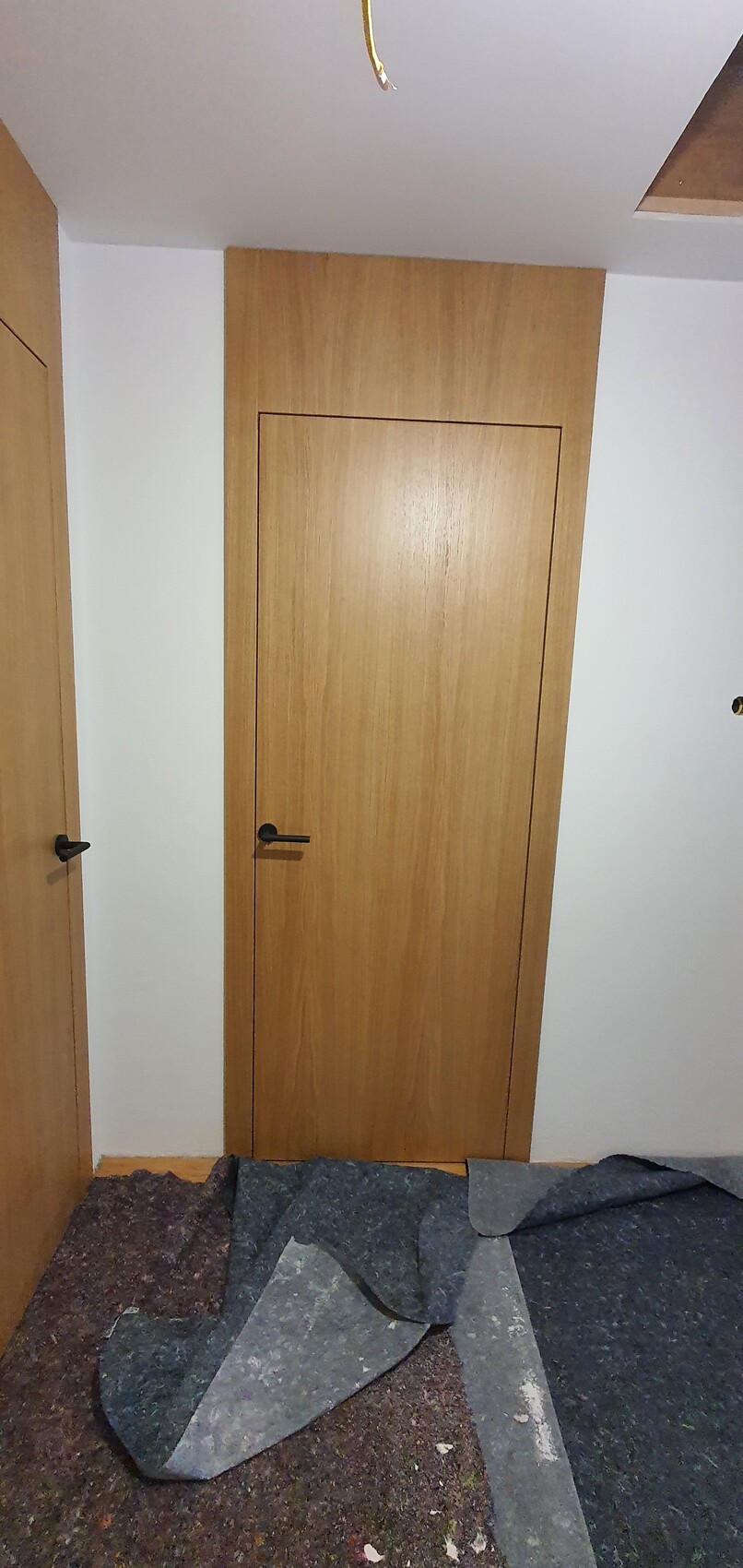 Holztür im Flur mit schwarzem Griff, weiße Wände, lose Teppichreste auf dem Boden.