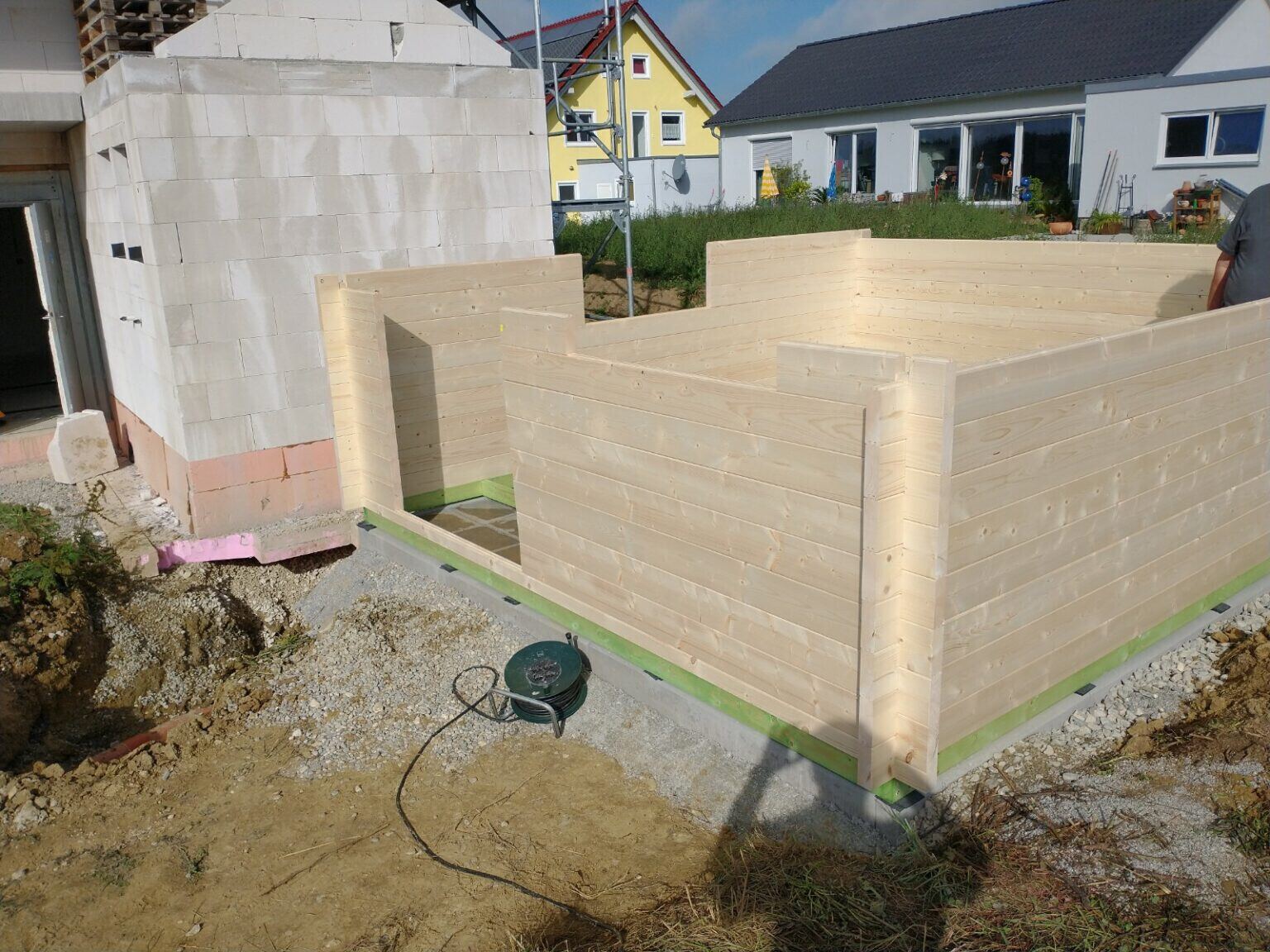 Neubau aus hellen Holzwänden mit Türöffnung, umgeben von Baugrund und Kies.