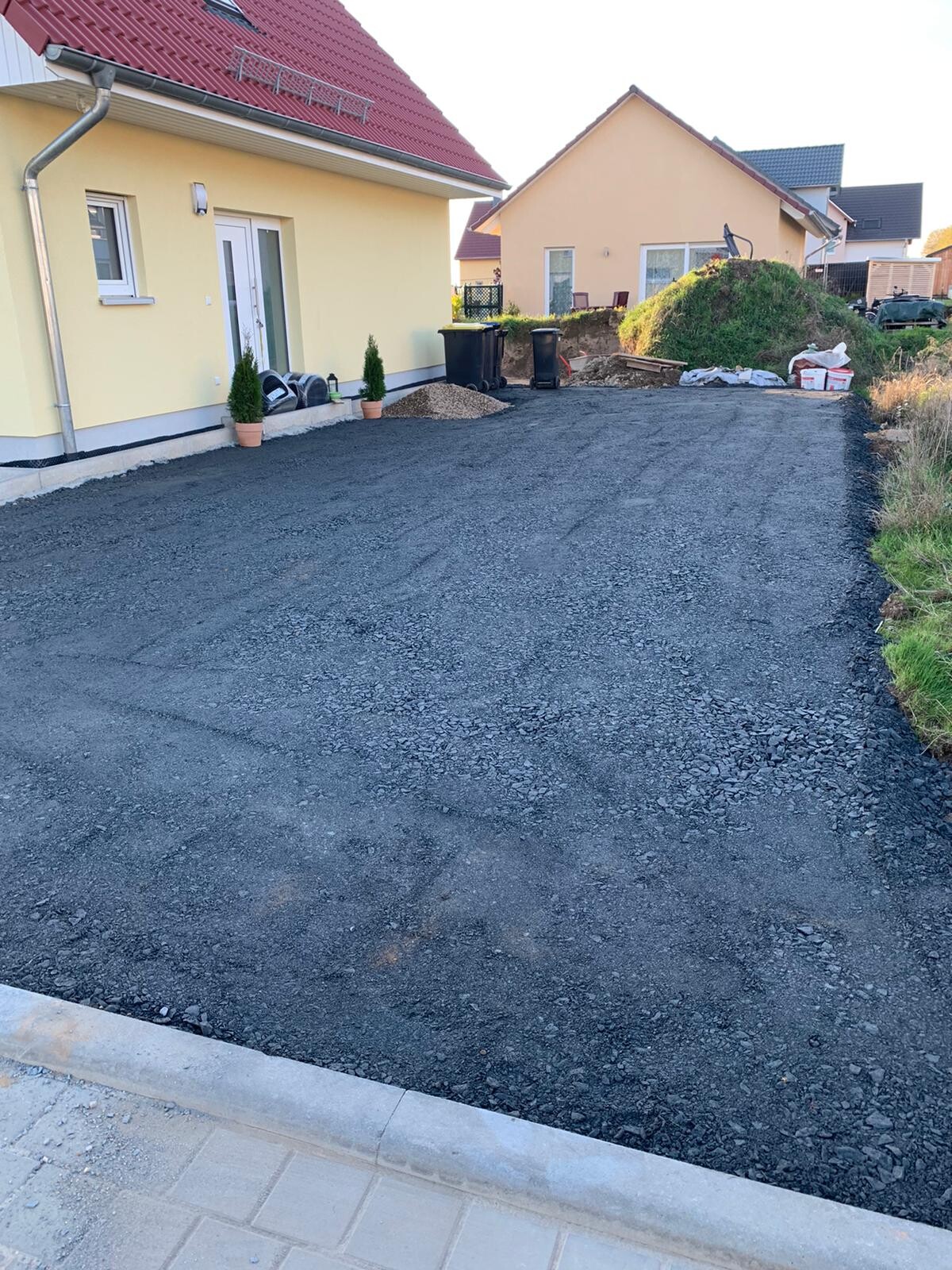 Neue Kiesauffahrt vor gelbem Einfamilienhaus; Gartengeräte, Mülltonnen und Kieshaufen im Hintergrund.