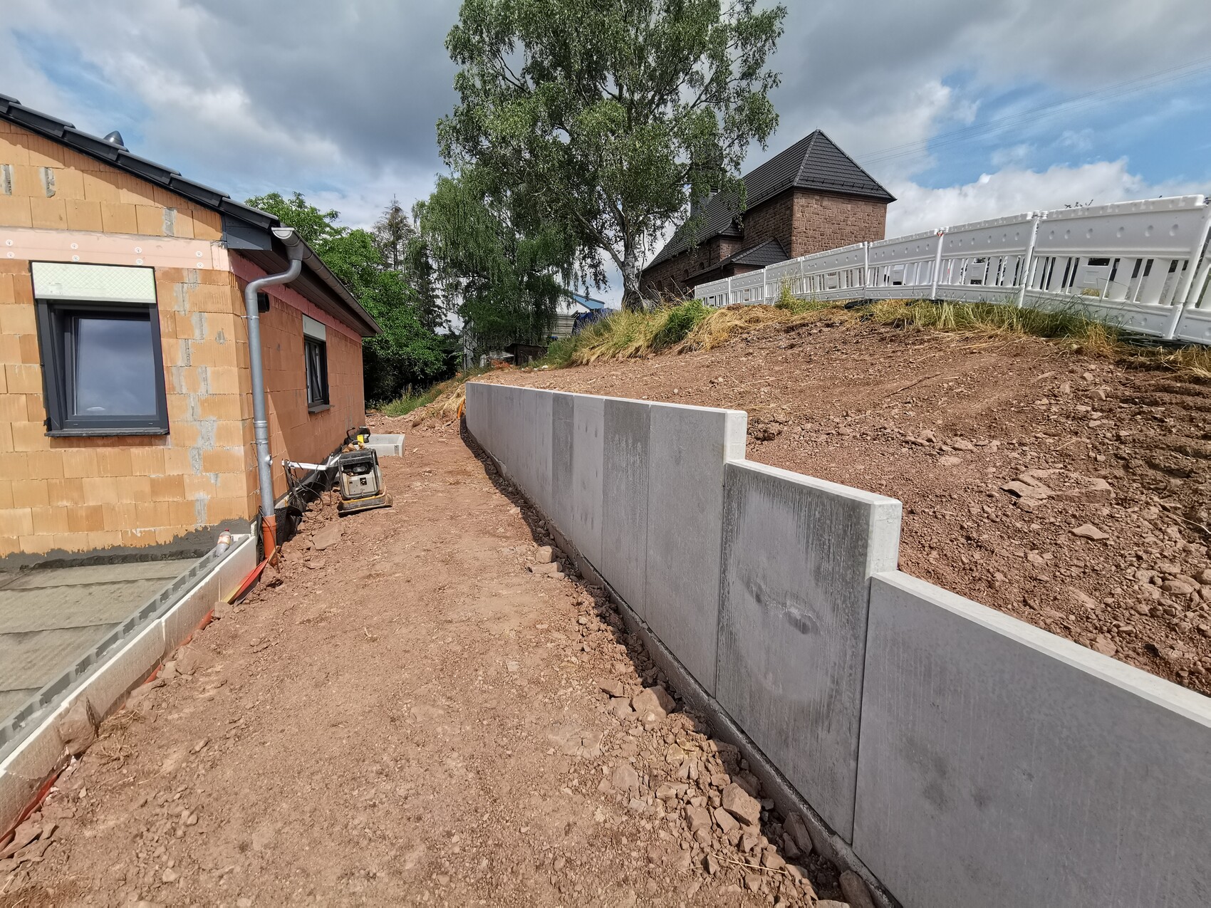 Baustelle am Hang: rotes Backsteinhaus, Betonmauer, Baugeräte und Bauzäune.