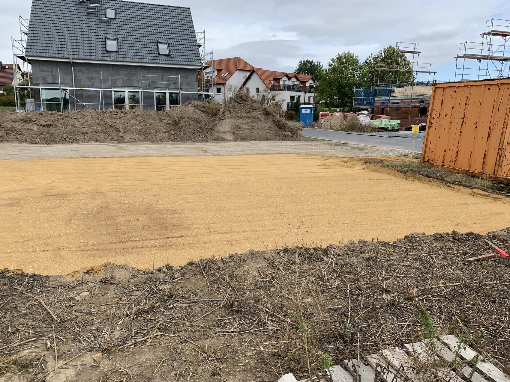 Baustelle mit neuem Haus, Gerüste, Sandboden, Schutt und oranges Baucontainer.