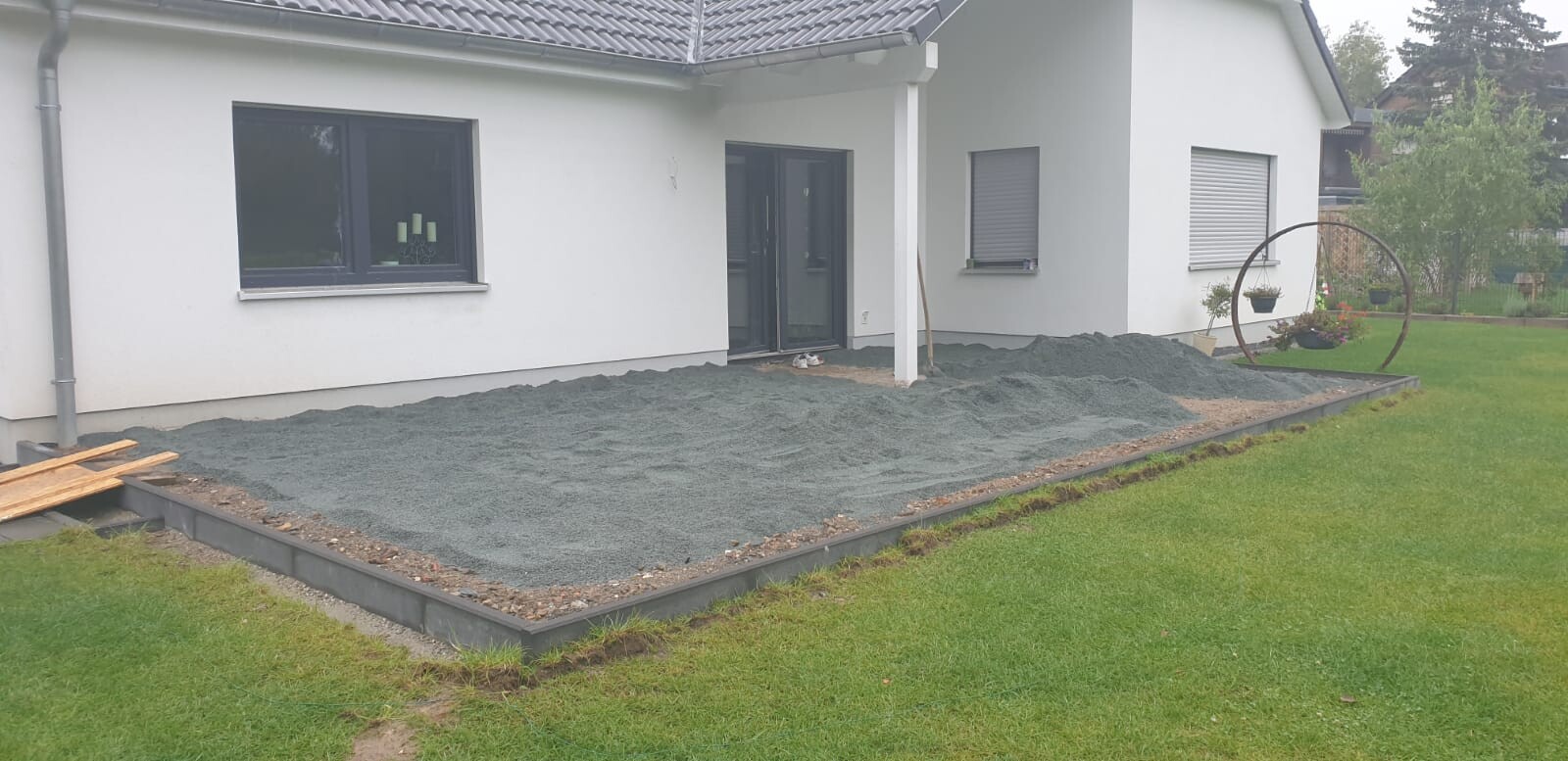 Grau geschotterte Terrasse vor weißem Wohnhaus; kreisförmiger Metallbogen mit Pflanzgefäßen.