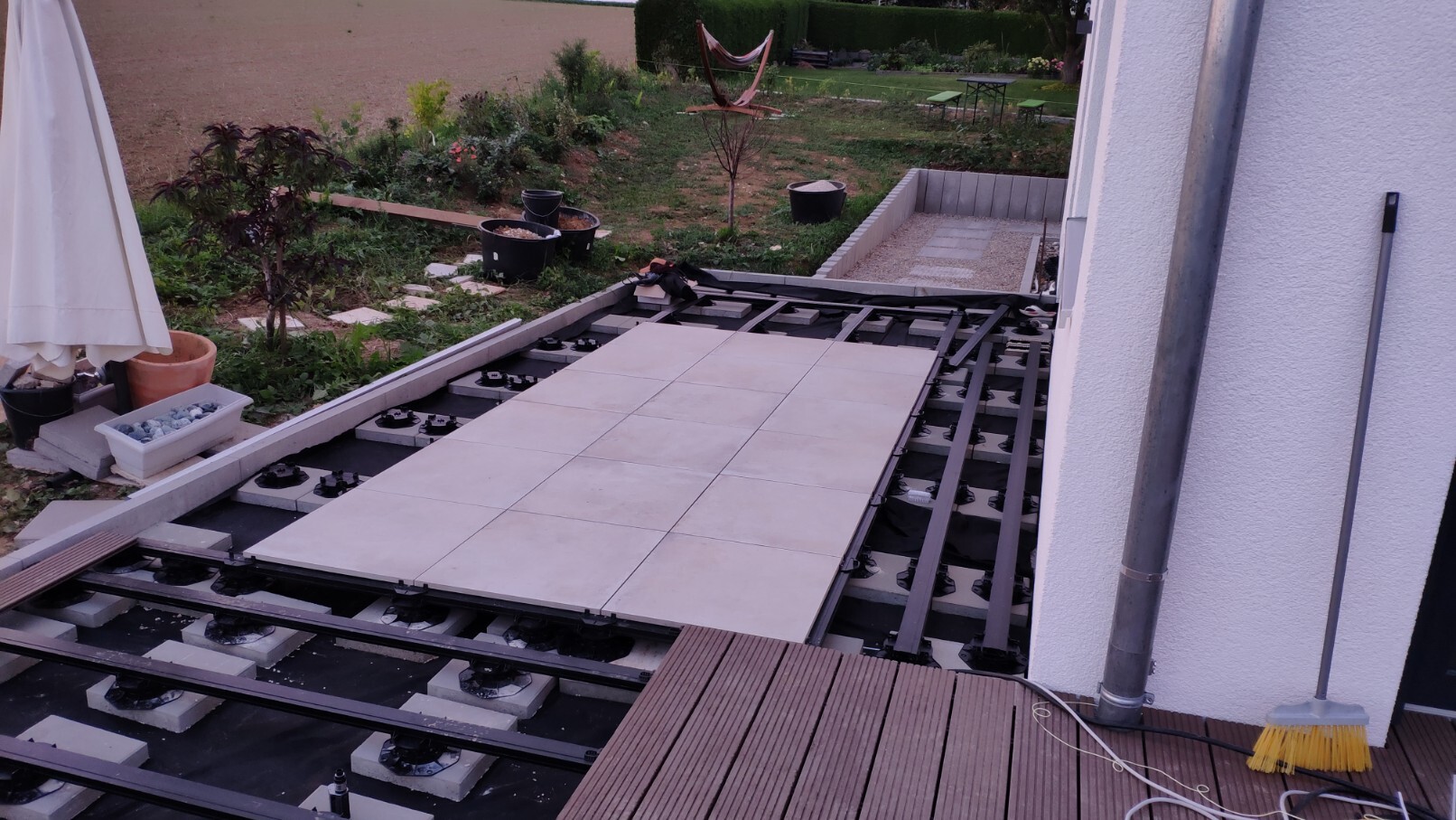 Außenbereich: Terrassenfliesen werden auf Unterkonstruktion verlegt; Garten mit Pflanzen im Hintergrund.