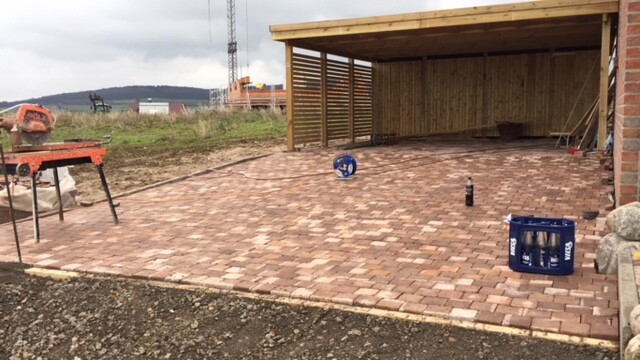 Gepflasterte Baufläche mit Holzcarport, linke Kreissäge, blaue Kiste in der Mitte.