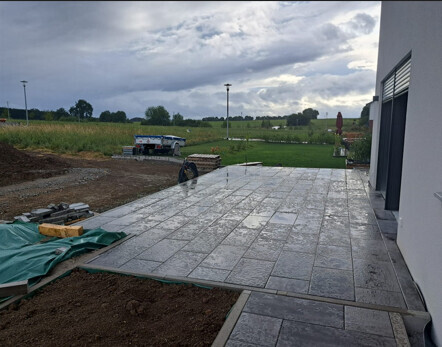 Neue gepflasterte Terrasse aus Steinplatten neben einem Gebäude, Baugeräte im Hintergrund.