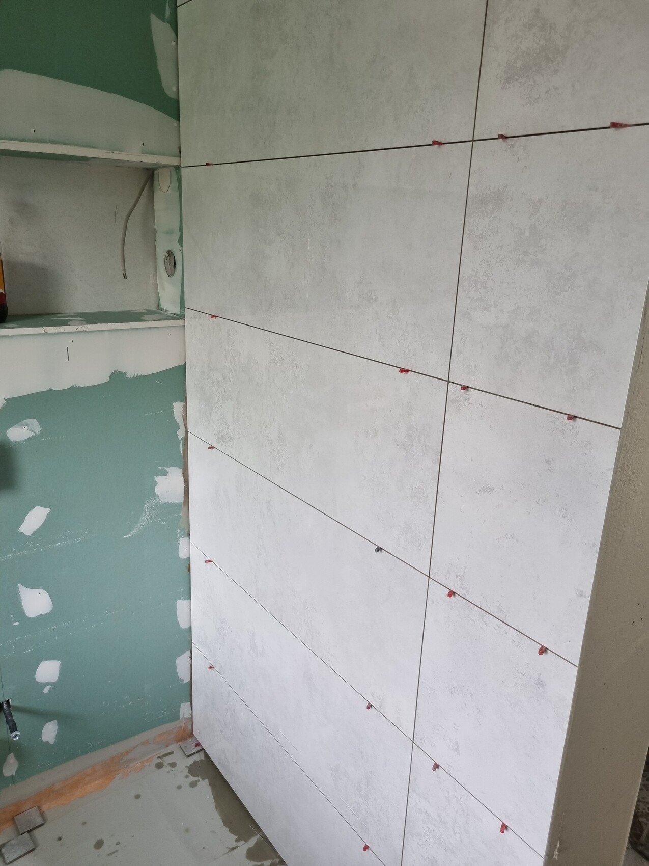 Großformatige weiß-graue Fliesen werden an einer Wand verlegt; rote Fliesenabstandhalter sichtbar.