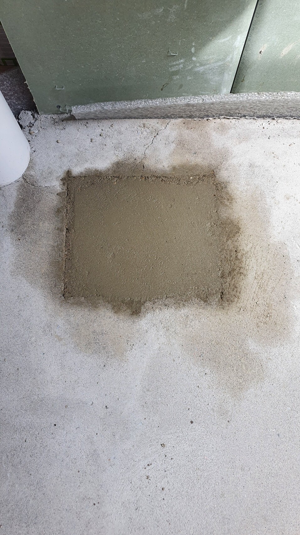 Frische Betonreparaturfläche auf dem Boden neben einem grünen Metallgehäuse; Rand leicht verlaufend.