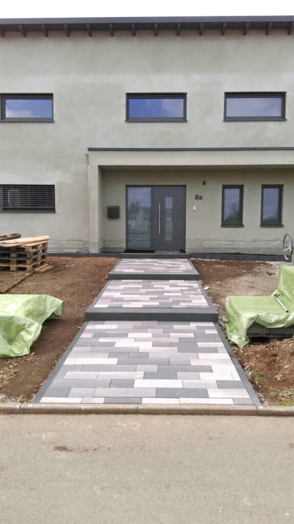 Moderne Gebäudefassade mit Eingang und gepflastertem grau-weißen Weg zur Tür, Baufläche links.
