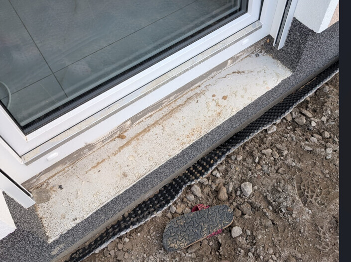 Fensterunterkante mit Betonrahmen, davor Erde und Kies, schwarze Drainageleiste am Boden.